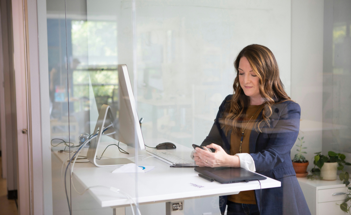 A woman who is sitting at a desk besides a computer screen and she's looking at her phone.