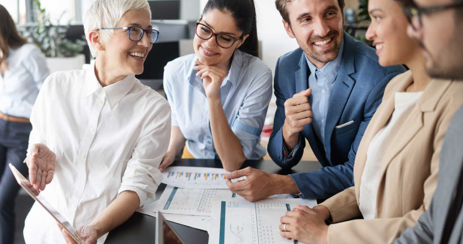 A group of business professionals standing around a raised desk, which has diagrams and charts on it.